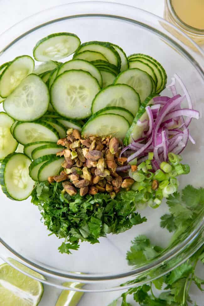 Clear glass bowl filled with sliced cucumber, red and green onion, cilantro and pistachios
