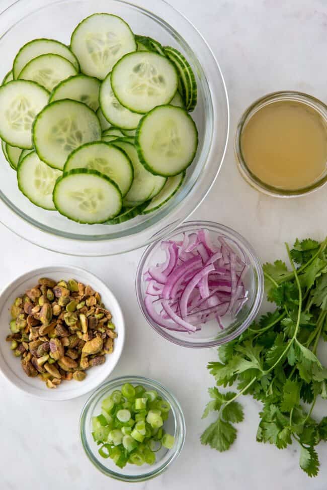 Glass bowls filled with sliced cucumber, red onion, green onion, and pistachios.