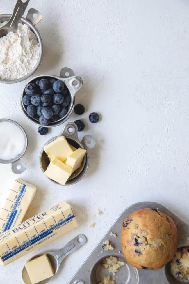 Vintage aluminum measuring cups filled with flour, butter, sugar and blueberries to learn how many tablespoons in a cup (tbsp to oz). A muffin tin sits next to them with a baked blueberry muffin.