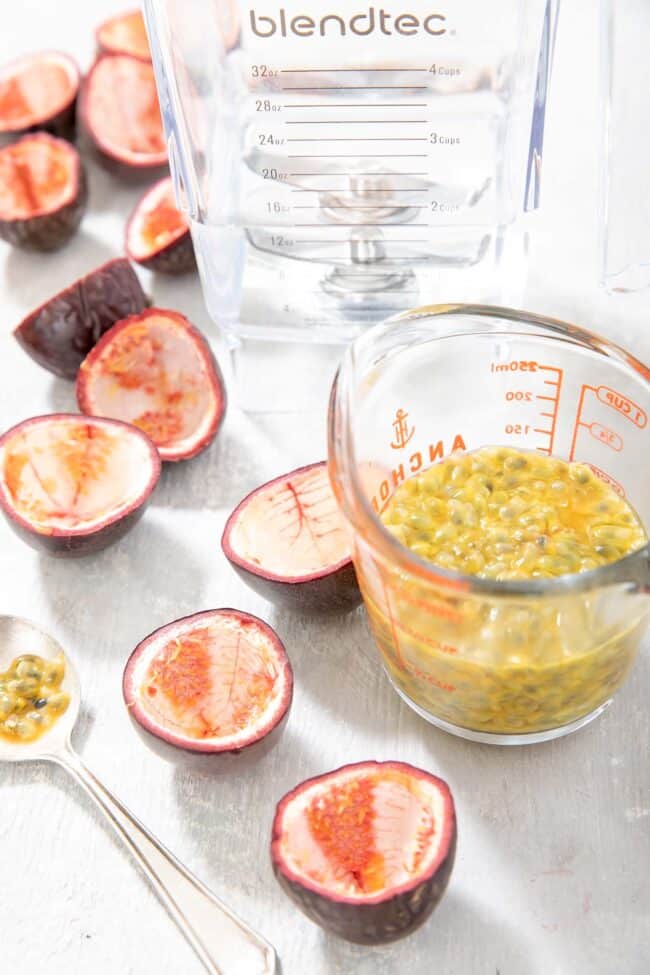 A glass measuring cup filled with passion fruit pulp. A spoon and cut open passion fruit sit next to the measuring cup.