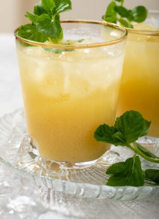 Two clear glasses with gold rims filled with fresh passion fruit juice, ice and mint sprigs.