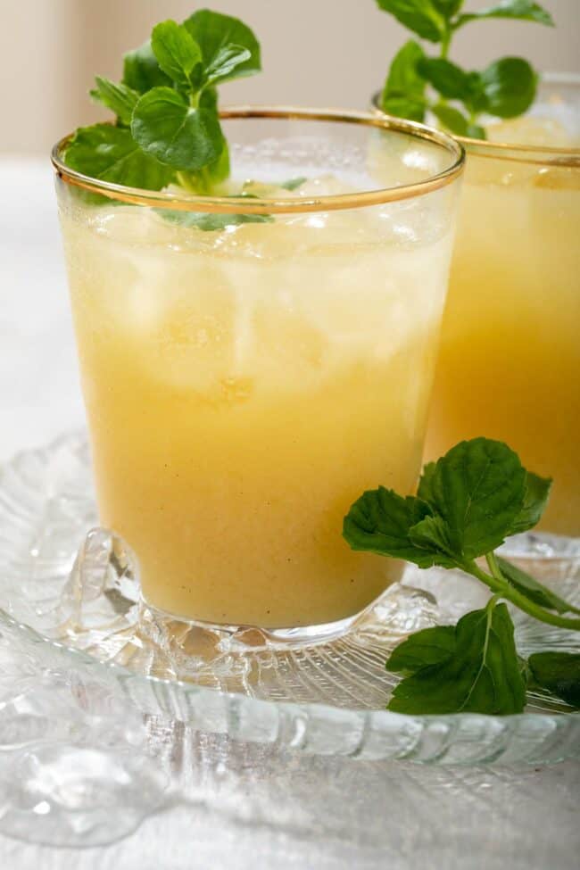 Two clear glasses with gold rims filled with fresh passion fruit juice, ice and mint sprigs.