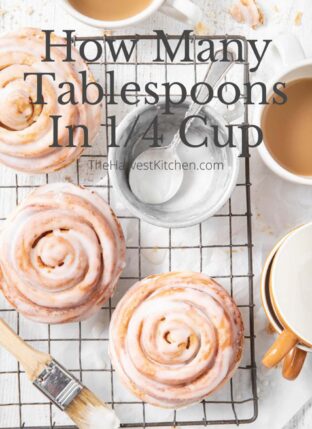 3 cinnamon rolls on a cooling rack with a measuring cup filled with icing - to measure how many tablespoons in 1/4 cup