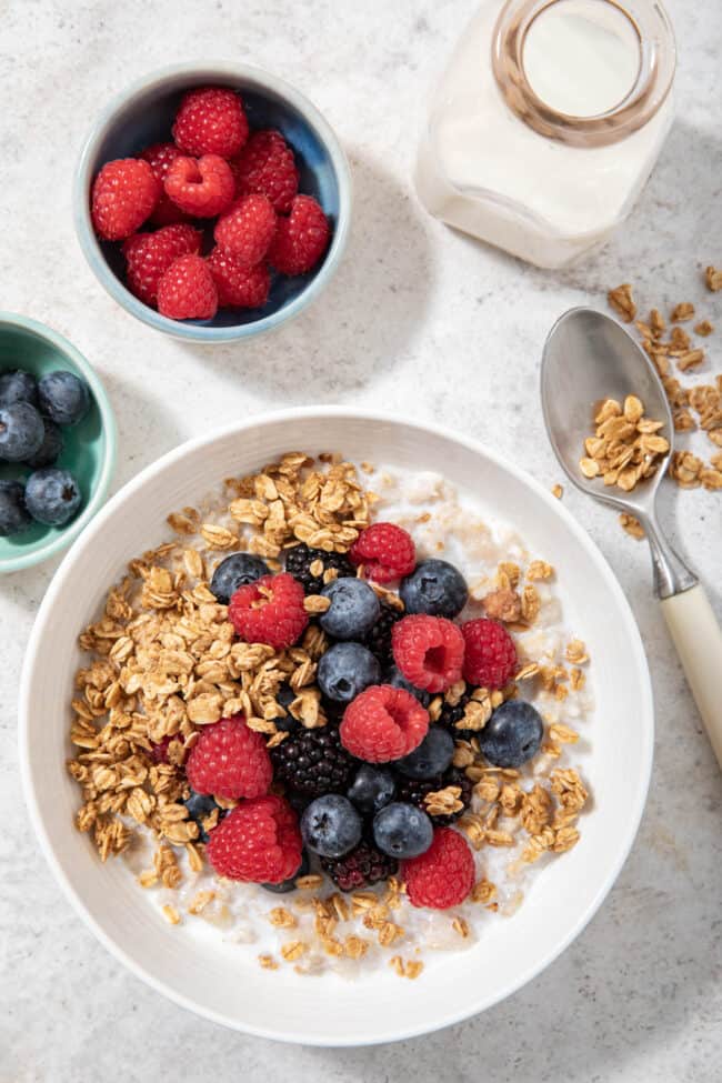 A white bowl filled with oatmeal and berries. A glass milk bottle and two small bowls filled with berries sits next to the white bowl.