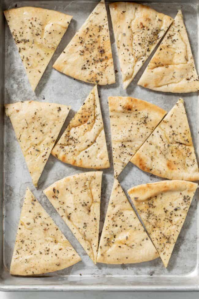 12 triangles of pita bread brushed with olive oil and sprinkled with dry herbs rest on a baking sheet.