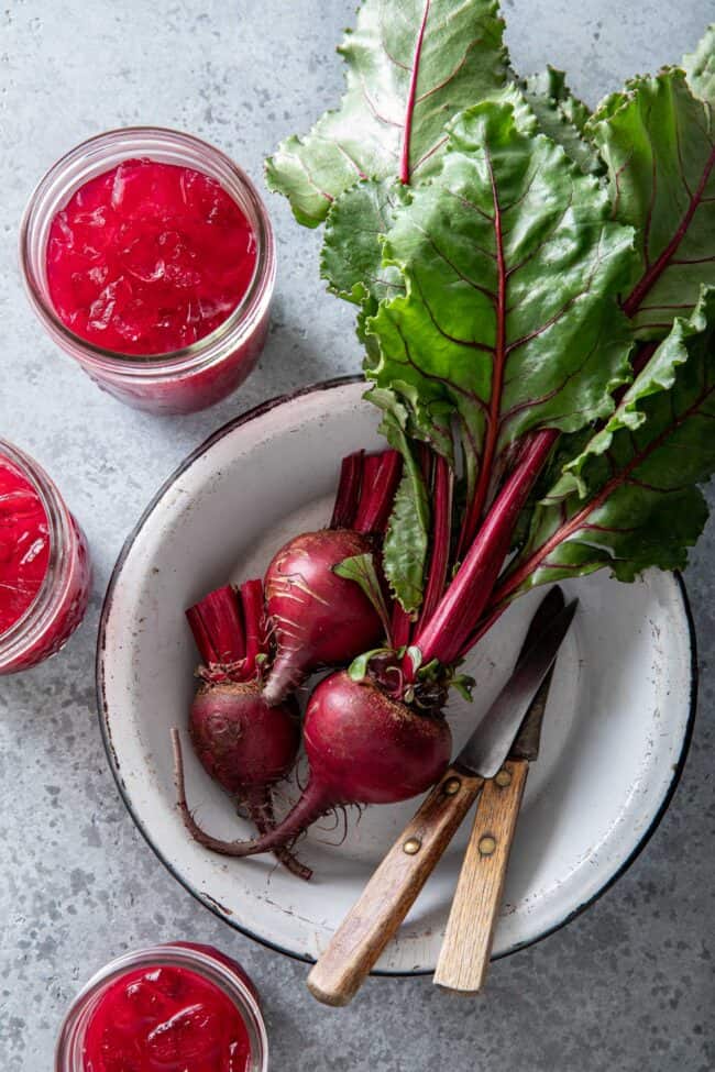 three beets with two knives in a white pie tin. Three glasses of beet juice sit next to the pie tin