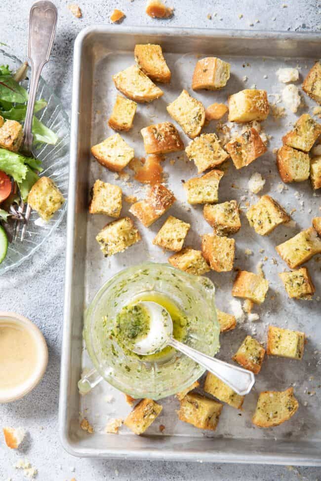 An aluminum cookie sheet filled with basil pesto croutons. A glass dish filled with salad and croutons sits next to the cookie sheet.
