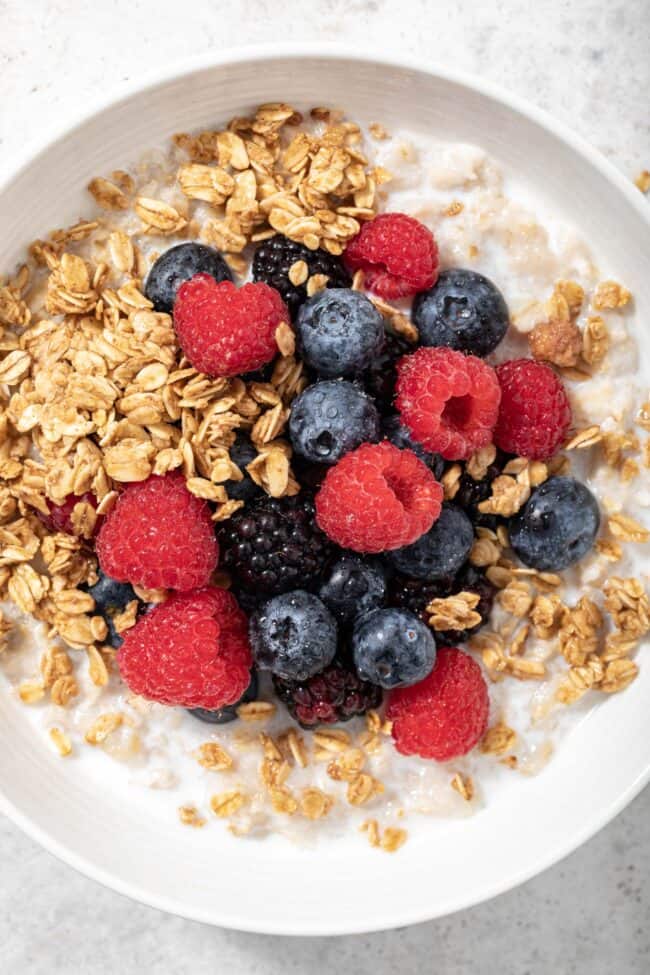 a white bowl filled with cooked oatmeal, sprinkled with granola, mixed berries and oat milk for article are oats gluten free