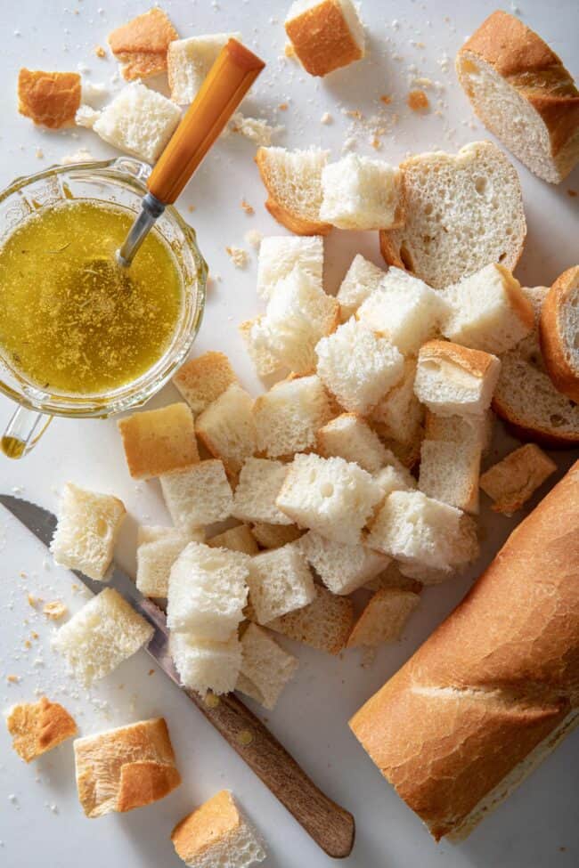 a French baguette cut into cubes sit next to a glass cup filled with melted butter.