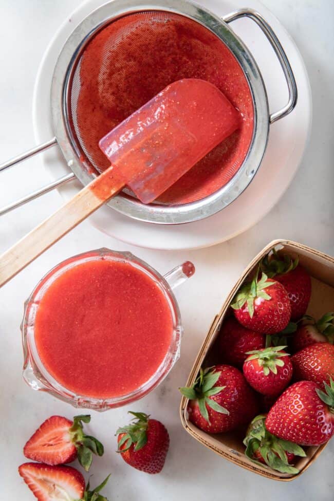 a glass measuring cup filled with strawberry puree sits next to a fine mesh strainer filled with the puree for making homemade strawberry lemonade