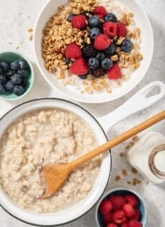 A white sauce pan filled with cooked oatmeal. A white bowl filled with oatmeal, granola and mixed berries sits next to the sauce pan.