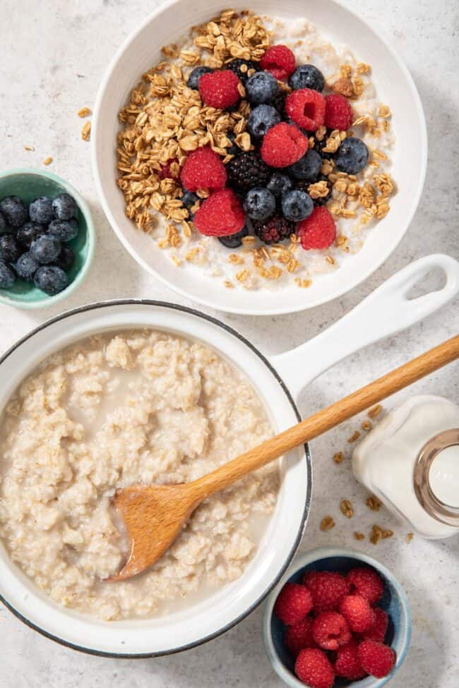 a white sauce pan filled with cooked oatmeal. A white bowl filled with oatmeal, granola and berries sits next to the sauce pan. for article are oats gluten free