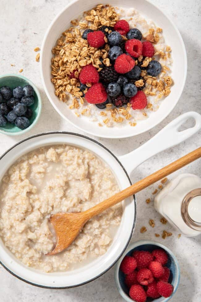 A white sauce pan filled with cooked oatmeal. A white bowl filled with oatmeal, granola and berries sits next to the sauce pan. for article are oats gluten free