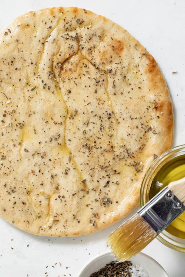 a flat pita bread brushed with olive oil and dried herbs. A pastry brush rests on a bowl of olive oil net to the bread.