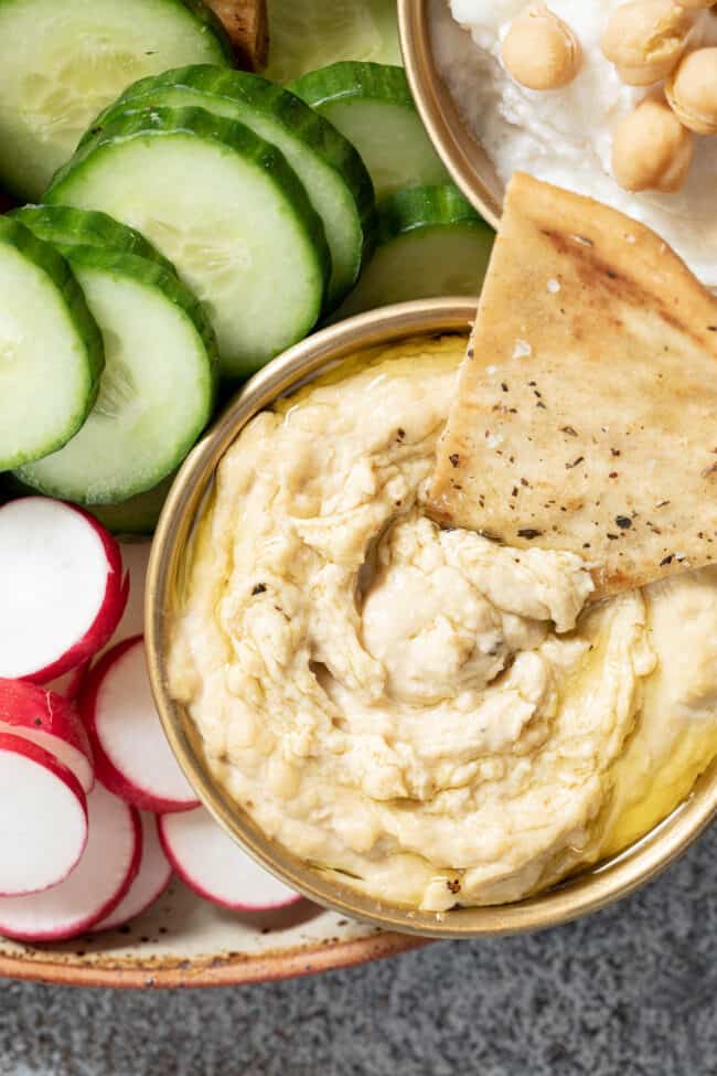 A bowl of hummus sitting on a plate next to slices of cucumber.