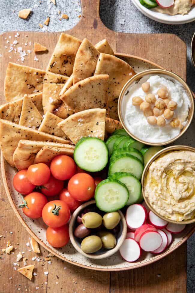 A plate filled with cherry tomatoes, sliced cucumber, two dips and homemade pita chips.
