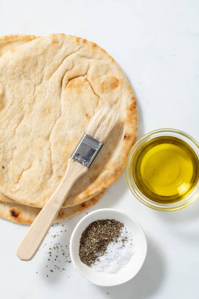 Pita bread sit next to a bowl of olive oil and a bowl of dried herbs - for how to make pita chips.