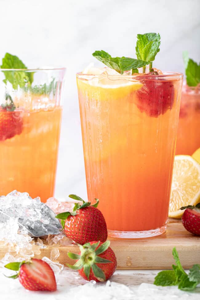 3 glasses filled with strawberry lemonade. Strawberries and crushed ice are scattered next to the glasses