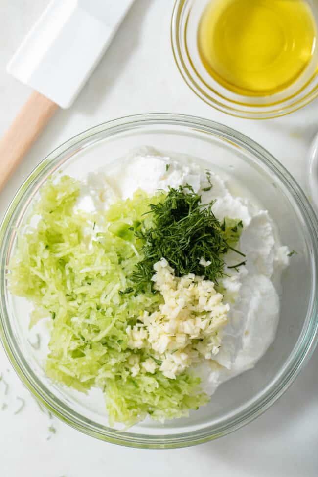 A clear glass bowl filled with Greek yogurt, grated cucumber, dill and garlic.