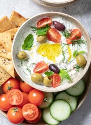 bowl filled with tzatziki sauce. A plate filled with pita chips, cherry tomatoes and slices of cucumber sit next to the bowl.