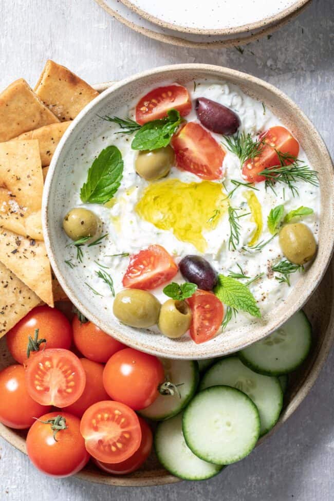 bowl filled with tzatziki sauce. A plate filled with pita chips, cherry tomatoes and slices of cucumber sit next to the bowl.