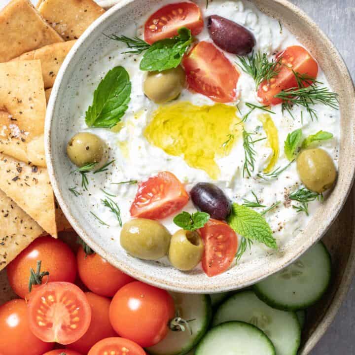 bowl filled with tzatziki sauce. A plate filled with pita chips, cherry tomatoes and slices of cucumber sit next to the bowl.