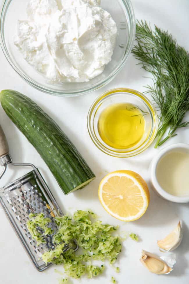 a clear glass bowl filled with yogurt, cucumber grated, a small bowl filled with lemon juice and oil - ingredients for learning how to make tzatziki
