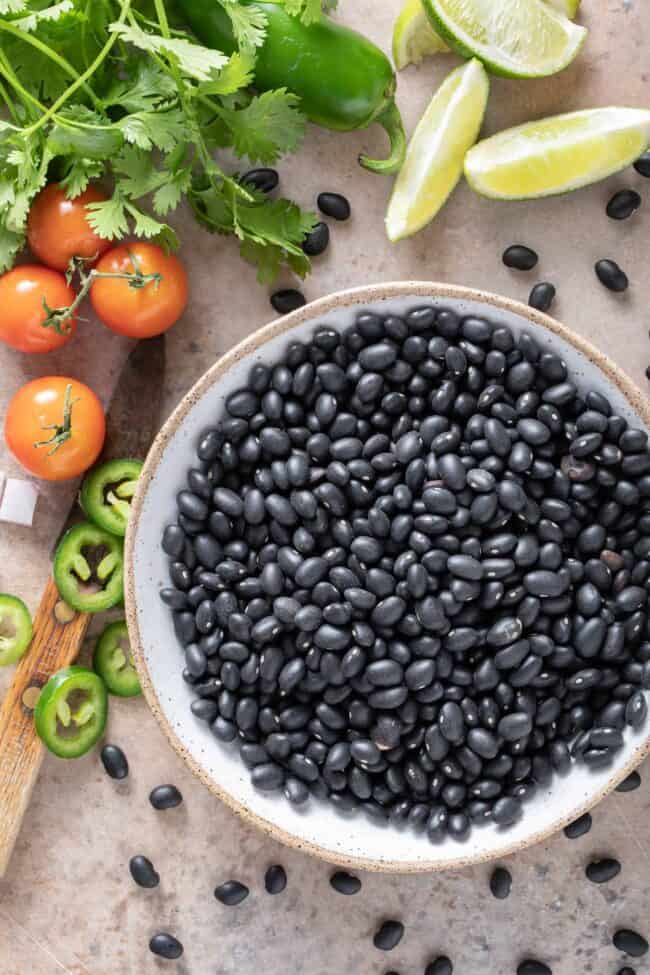 A white bowl filled with legumes. Cilantro, cherry tomatoes and slices of jalapeno sit next to the bowl.