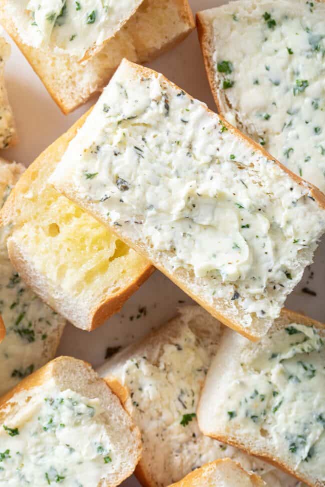 Several pieces of cut baguette are scattered on a white cutting board. The bread slices are slathered generously with garlic and herb butter for Italian garlic bread