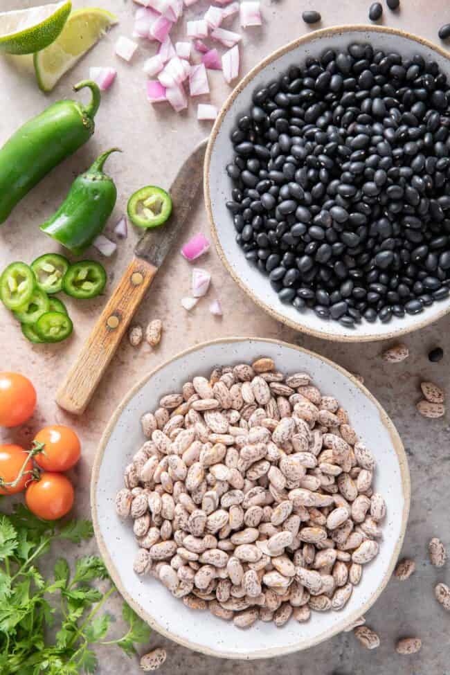 Two bowls sitting on a table - one filled with uncooked black beans and the other with uncooked pinto beans. A knife, tomatoes, jalapenos, cilantro and onions sit next to the bowls.