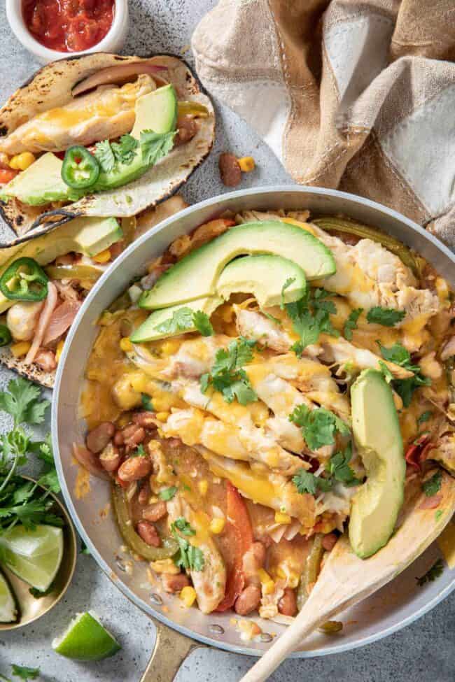 A metal skillet filled with chicken fajita casserole and avocado slices. A wooden spoon rests in the skillet.