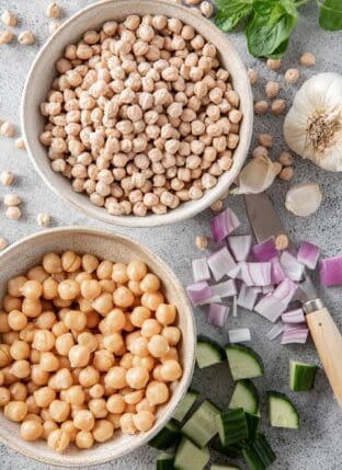 Two bowls filled with chickpeas - one cooked and one dried. Chopped cucumber and red onion and a knife sit next to the bowl for chickpeas vs garbanzo beans