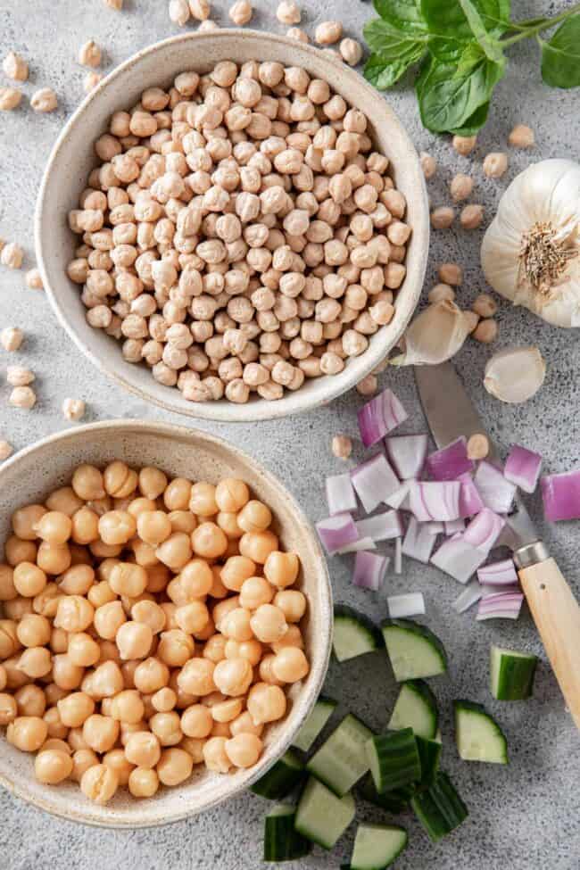 Two bowls filled with chickpeas - one cooked chickpeas and one raw. Chopped cucumber, purple onion, fresh mint and a knife sits next to the vegetables. For chickpeas vs garbanzo beans.