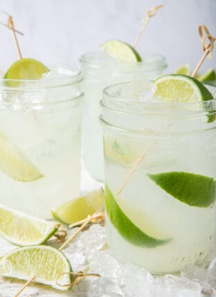 Three clear drinking glasses filled with limeade, ice and lime wedges.