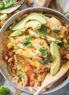 A metal skillet filled with chicken fajita casserole and avocado slices. A wooden spoon rests in the skillet.