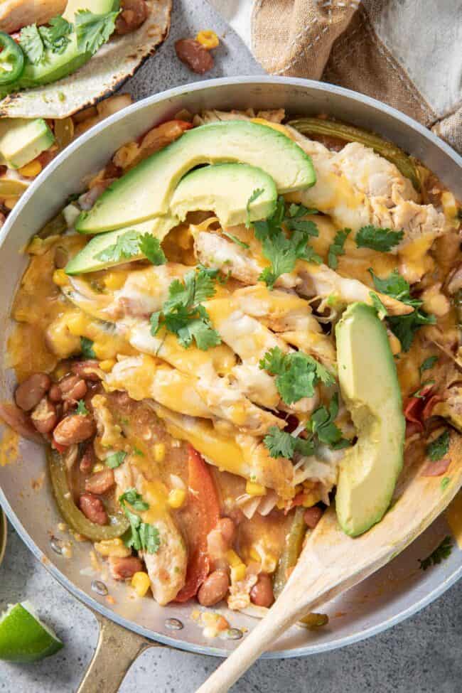 a metal skillet filled with chicken fajita casserole and avocado slices. A wooden spoon rests in the skillet.