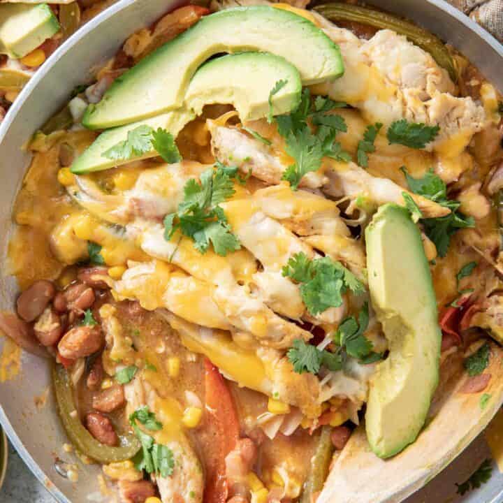 A metal skillet filled with chicken fajita casserole and avocado slices. A wooden spoon rests in the skillet.