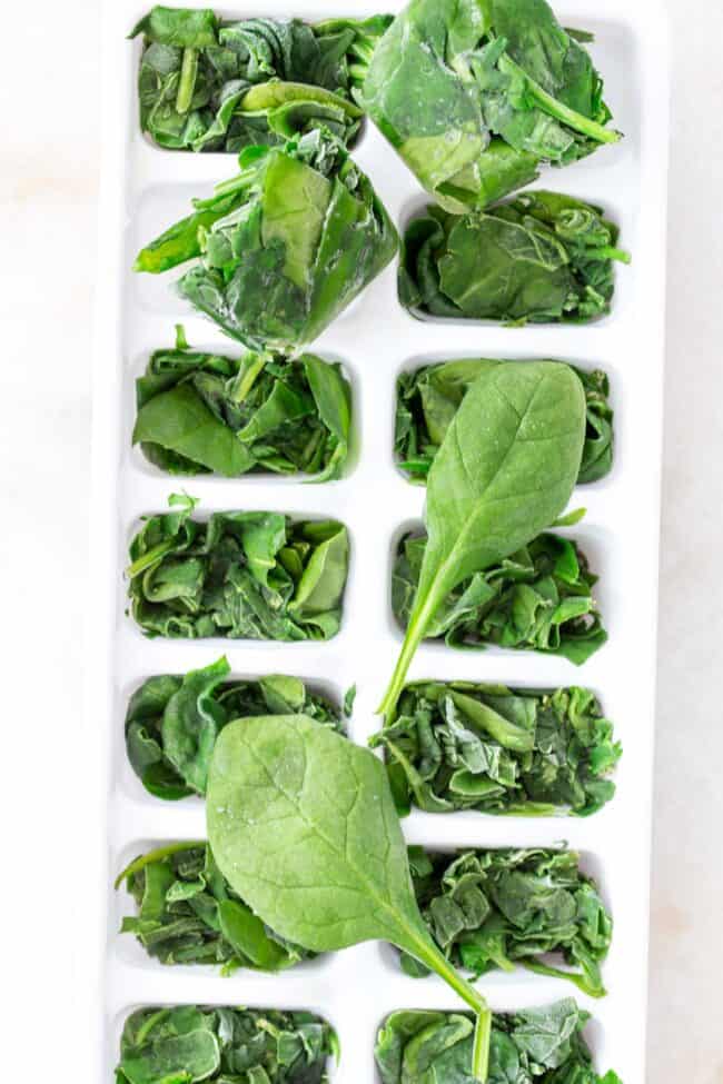 A white ice cube tray filled with green vegetable leaves. 