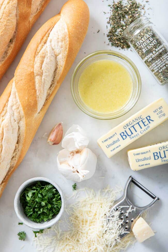 Two French baguettes sit next to a bowl of melted butter, two sticks of butter, a jar of Italian herbs, a small bowl of chopped parsley and garlic and grated parmesan cheese.