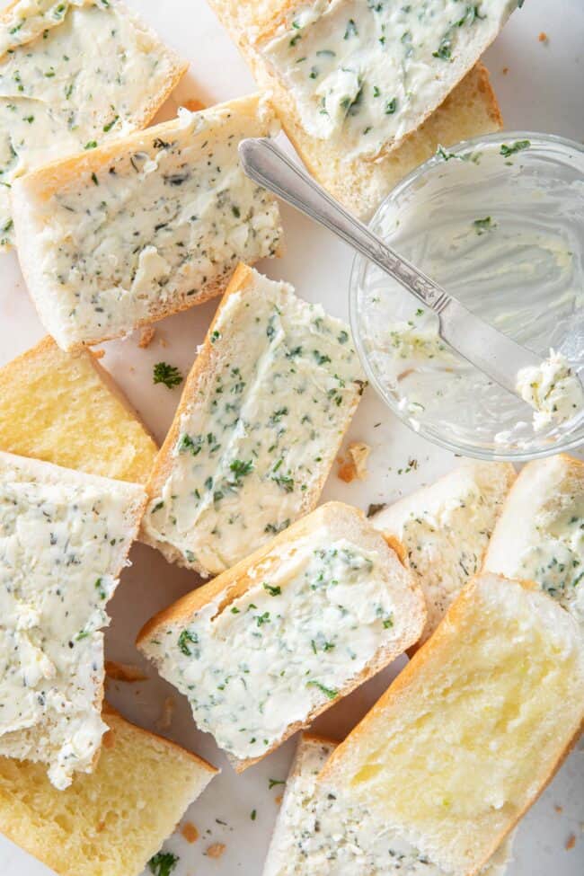 Several cut pieces of baguette are slathered with garlic and herbed butter. A clear bowl with a little smeared butter in it and a small butter knife sit next to the pieces of bread.