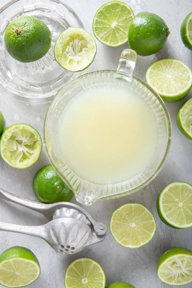 A glass measuring cup filled with lime juice. Limes sliced in half and a metal reamer sit next to the measuring cup.