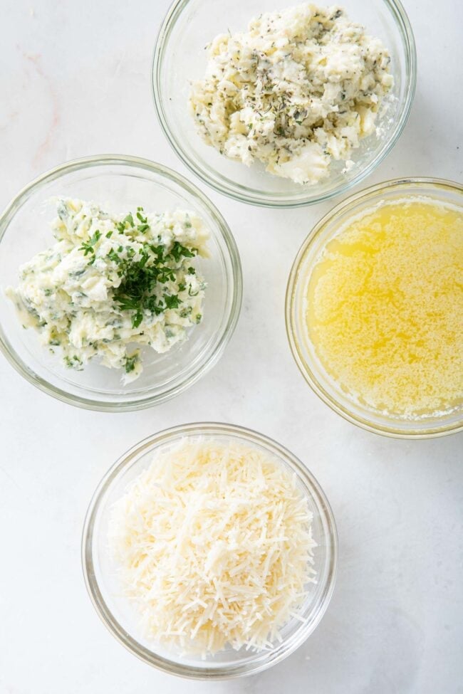 Four clear glass bowls, one filled with mashed garlic and herb butter, one with mashed parsley butter, one with melted butter and one with grated parmesan cheese.