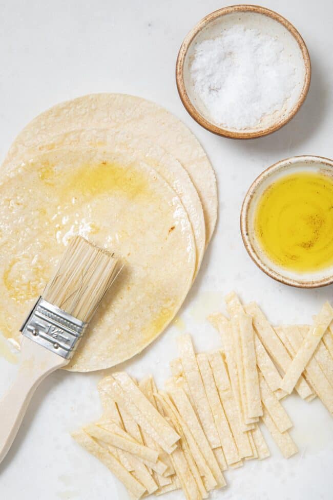 corn tortillas on a cutting board. A small bowl filled with olive oil sits next to a brush for brushing the tortillas with.