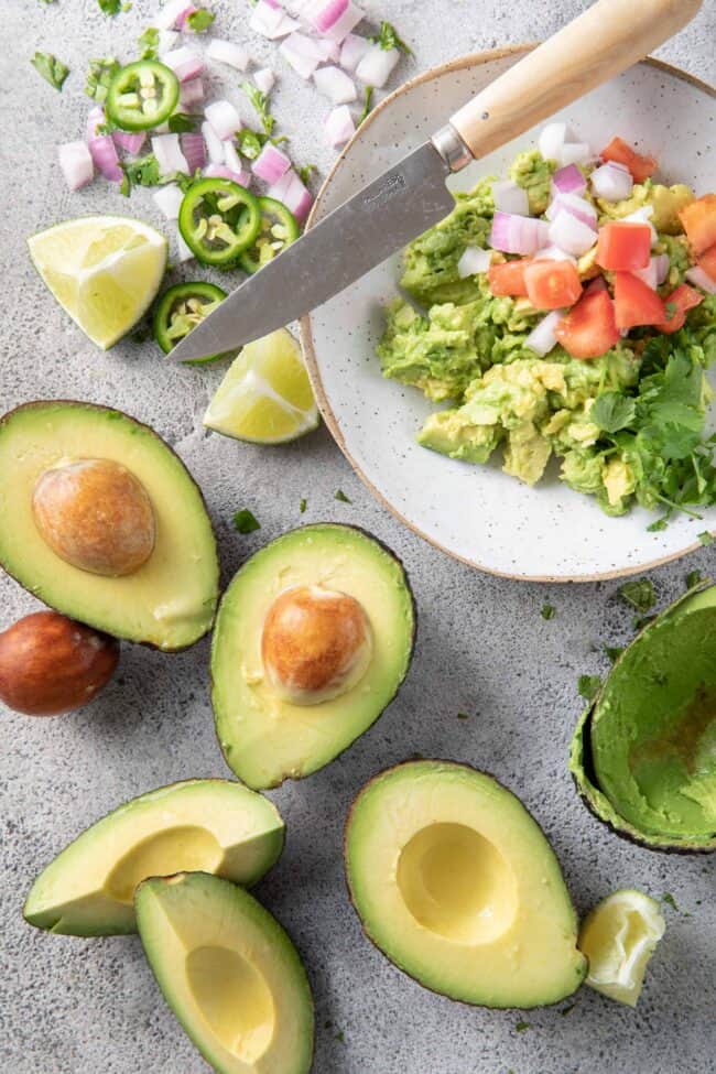 Several avocados are sliced in half with a bowl of mashed fruit sitting next to them
