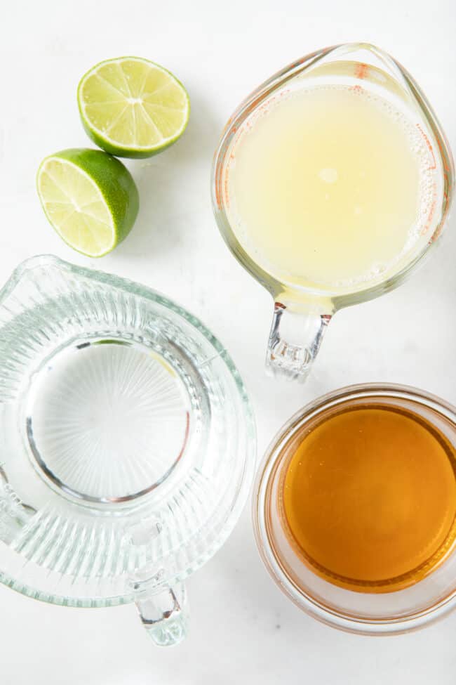 A clear glass measuring cup filled with water, another clear glass measuring cup filled with lime juice and a clear glass measuring cup filled with honey.