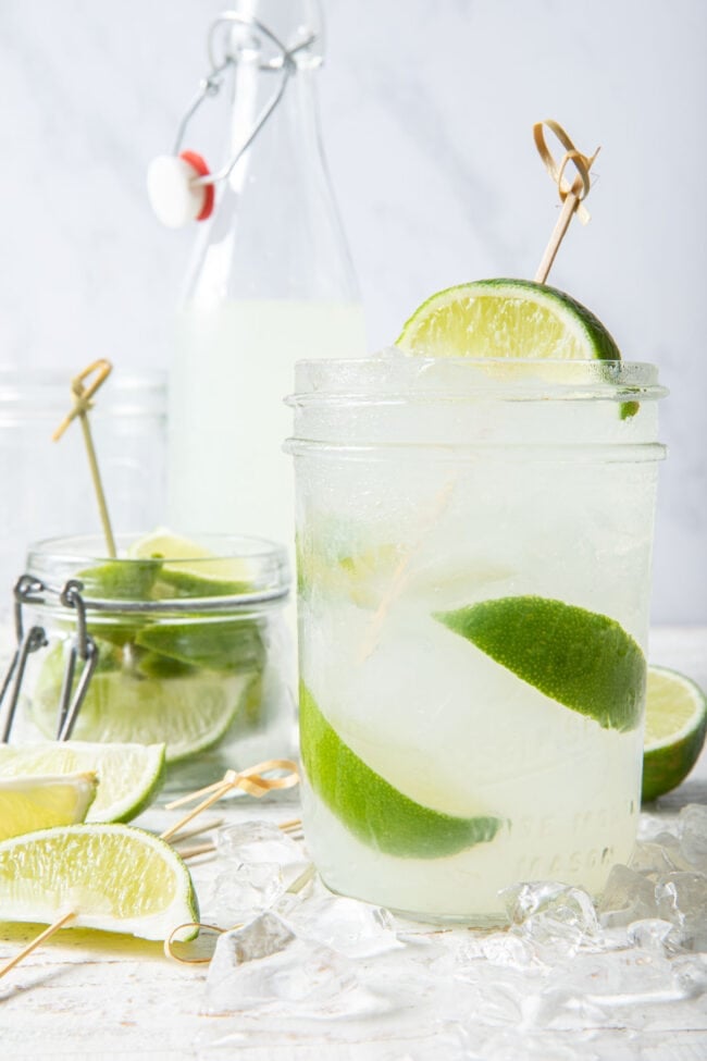 A clear drinking glass filled with clear liquid and ice. A small mason jar filled with slices of citrus sits next to the glass.