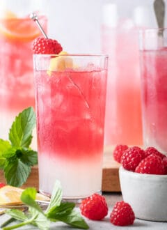 Four glasses filled with pink lemonade. A small bowl of raspberries sits next to the glasses.