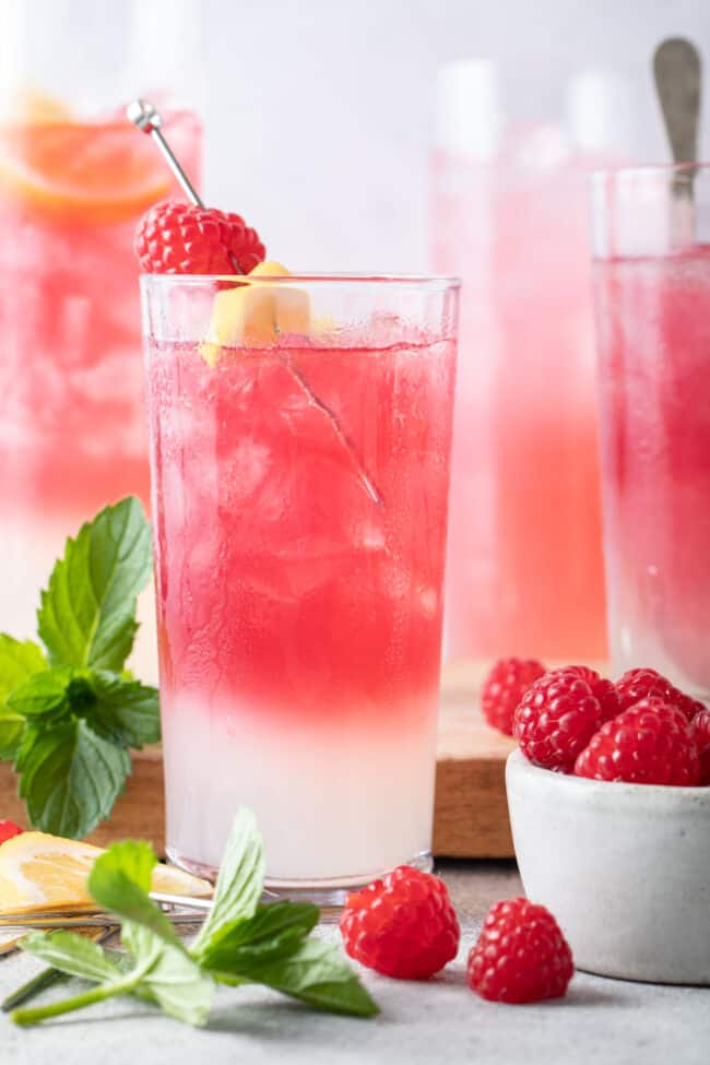 Four clear glasses filled with pink lemonade. A small bowl filled with raspberries sits next to the glasses.