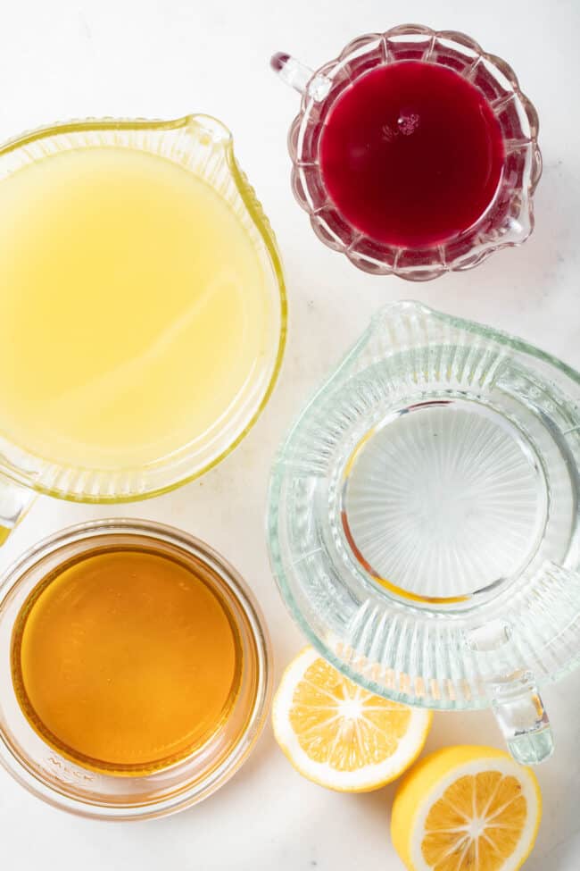 Eight clear glass liquid measuring cups filled with lemon juice, water, honey and cranberry juice.