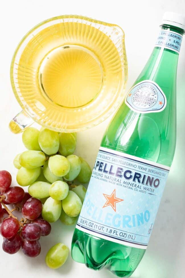 A bottle of Pellegrino sparkling water sits next to a measuring cup of white grape juice and red and green grapes to make a non alcoholic spritzer